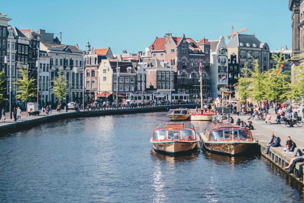 Twee rondvaartboten liggen afgemeerd in een Amsterdamse gracht, met toeristen op de kade en historische gevels erachter